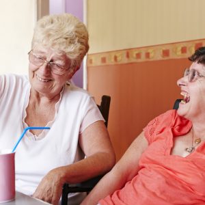 Two women laughing. One woman is pouring tea into a mug with a straw for the other woman, who is in a motorised wheelchair.