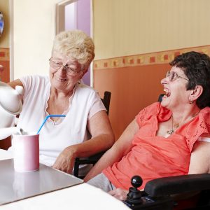 Two women laughing. One woman is pouring tea into a mug with a straw for the other woman, who is in a motorised wheelchair.