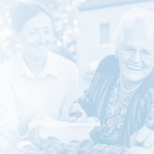 An older woman with white hair and a younger woman with brown hair at an outdoor produce stand. The older woman is reaching forward to select a piece of fruit. The younger woman is holding a bag open for her. Both are smiling.