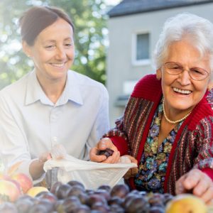 An older woman with white hair and a younger woman with brown hair at an outdoor produce stand. The older woman is reaching forward to select a piece of fruit. The younger woman is holding a bag open for her. Both are smiling.